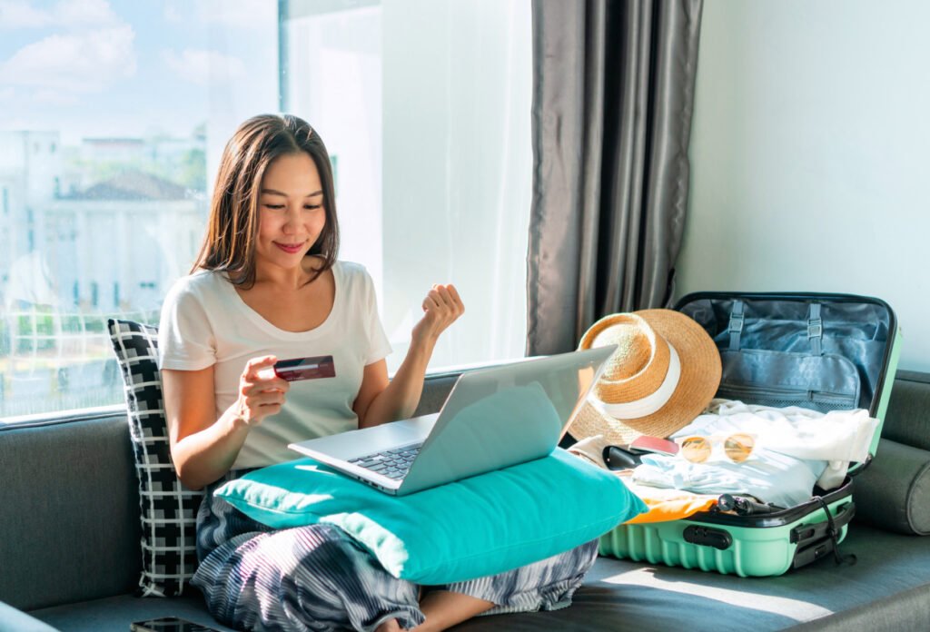 Happy woman using credit card for booking flight tickets and hotel at home with luggage packed beside her